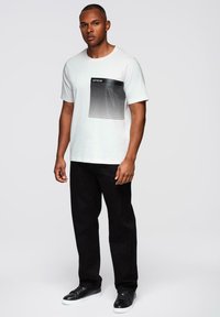 Bílá tričko s krátkým rukávem s černým obdélníkovým designem s vodorovnými pruhy a textem. V kombinaci s černými kalhotami a černými teniskami.