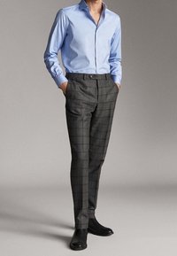 Man in een lichtblauw overhemd, met grijs geruite broek en zwarte laarzen, staand met de handen in de zakken tegen een effen achtergrond.
