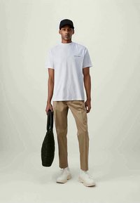T-shirt bianca a maniche corte con un piccolo logo, abbinata a pantaloni beige e una borsa nera. Il modello indossa un cappello nero e scarpe da ginnastica bianche.
