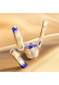 Shiseido Vital Perfection hudpleieprodukter i gull- og lilla emballasje: to serum og en krem, plassert på en teksturert gullbakgrunn.