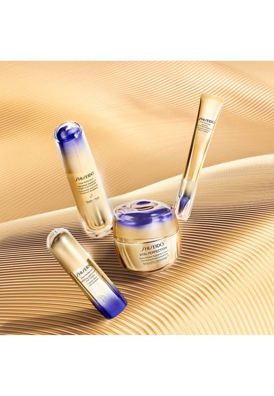 Productos de cuidado de la piel Shiseido Vital Perfection en envases dorados y morados: dos sérums y una crema, colocados sobre un fondo texturizado dorado.