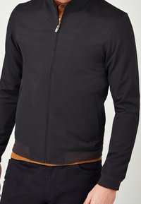 Zwarte jas met ritssluiting en opstaande kraag, voorzien van zijzakken en een geribde zoom. De binnenkant is afgewerkt met een bruine voering.