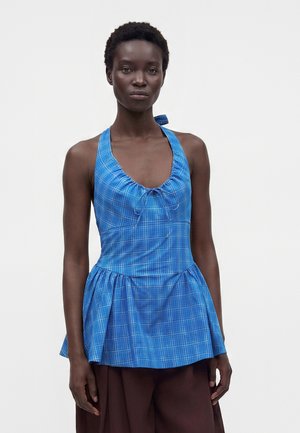 Femme portant un haut à carreaux bleu à col halter avec un ourlet peplum et un pantalon marron foncé sur un fond clair uni.