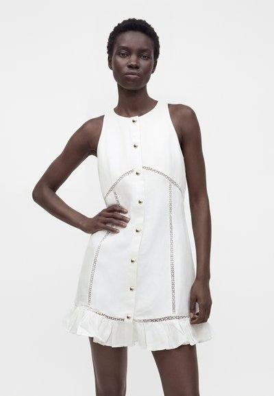Femme noire portant une robe blanche sans manches avec des boutons dorés, des détails en dentelle et un ourlet volanté, debout avec une main sur la hanche.
