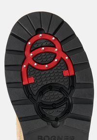 Černá gumová podrážka s červenými kruhovými akcenty a cvočky, s texturovaným designem a drážkami pro lepší přilnavost. Viditelné jméno značky.