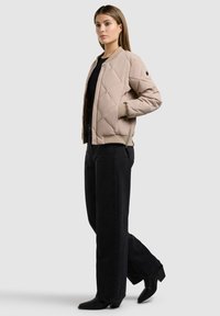 Jeune femme portant une veste bomber matelassée beige, un chemisier noir, un pantalon large noir et des bottines noires, debout de profil sur un fond gris.
