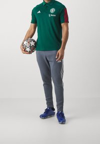 Zelené polo tričko s logem Manchester United a červenými pruhy, spárované se šedými tepláky a modrými kopačkami. Drží černobílý fotbalový míč.