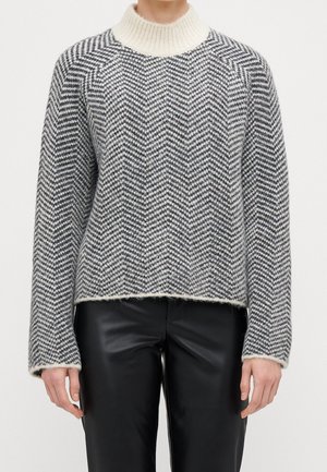 Personne portant un pull gris et blanc à motif chevron avec un col roulé crème et un pantalon en cuir noir, debout devant un fond uni.