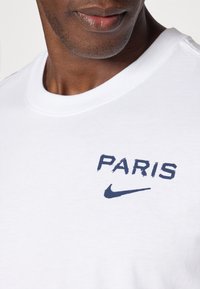 Hvid bomulds T-shirt med navy "PARIS" tekst og Nike swoosh-logo. Blød tekstur og klassisk rund hals design.
