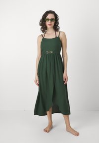 Vestido verde escuro, sem mangas, com corpete ajustado, cinto na cintura com metal decorativo e bainha assimétrica.