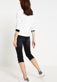 Sudadera blanca de manga larga con puños negros, combinada con jeans negros recortados y zapatillas blancas con detalles grises. Vista posterior.