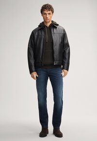 Zwarte leren jacket over een donkergroene gebreide hoodie, gecombineerd met een spijkerbroek en bruine suède laarzen, staande op een neutrale achtergrond.