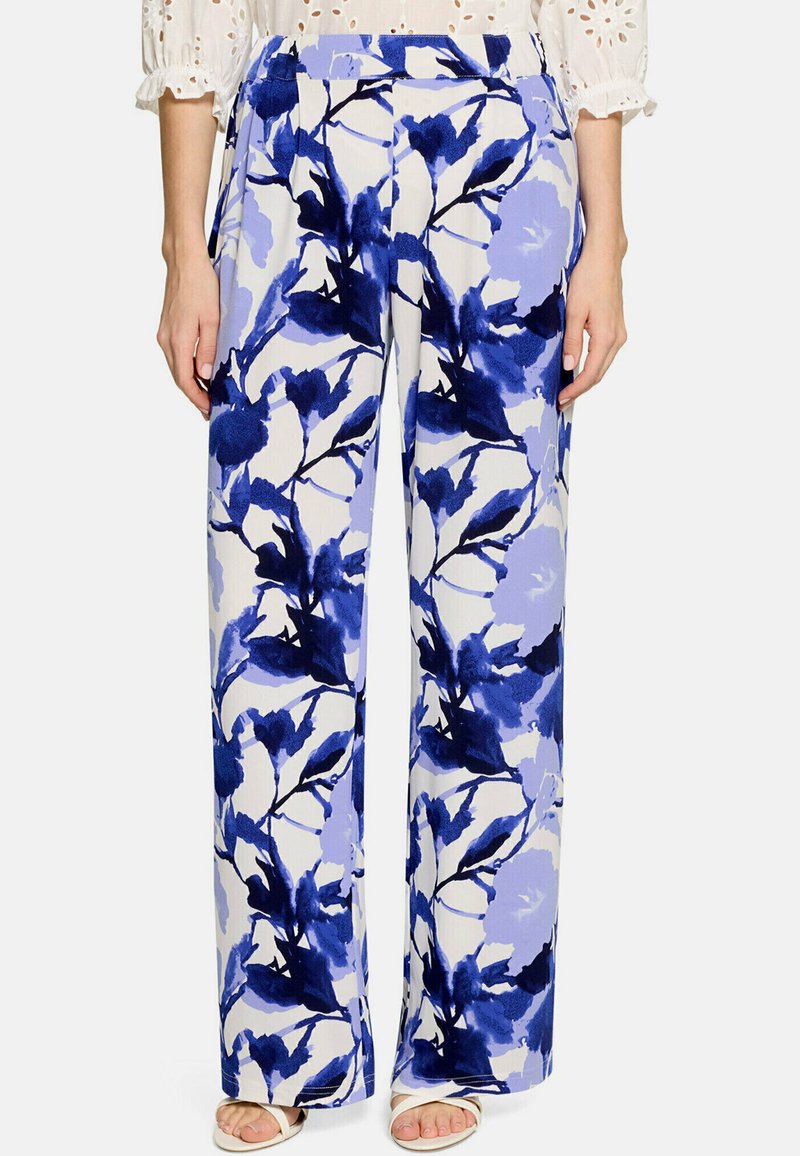 Pantalon blanc à jambes larges avec un imprimé floral bleu vif, porté avec des sandales blanches et un chemisier blanc à œillets.