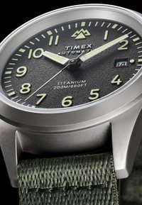 Titaniumur med tekstureret sort urskive, lysende tal og visere, datovindue, grøn stofrem og sølvkasse. Vandtæt ned til 200 meter.