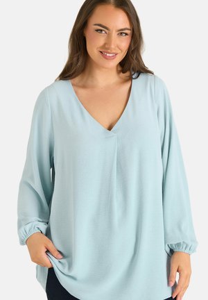 Kobieta z długimi brązowymi włosami, ubrana w jasnoniebieską bluzkę z dekoltem w serek i długimi rękawami, uśmiechająca się i trzymająca brzeg bluzki jedną ręką.