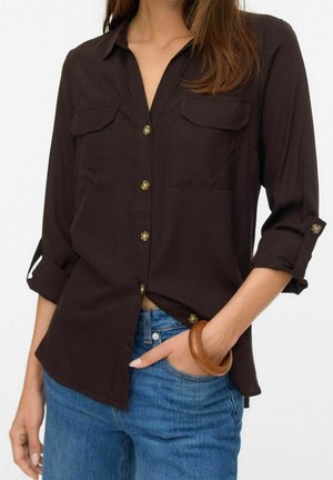 Femme portant une chemise marron foncé boutonnée aux manches retroussées et un jean bleu, avec une main dans la poche et un bracelet en bois au poignet.