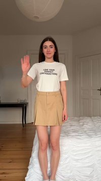Jeune femme debout dans une chambre, faisant signe de la main, portant une chemise blanche avec le texte « AIME TES CONTRADICTIONS PARFAITES » et une jupe beige.