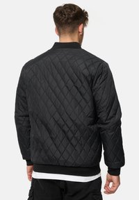 Veste noire matelassée avec un motif en losange, dotée d'un col et de poignets côtelés, ainsi qu'une texture lisse et brillante à l'extérieur.