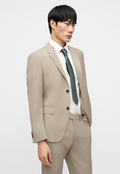 Μπεζ κοστούμι ραμμένο με υφή, που περιλαμβάνει σακάκι με δύο κουμπιά, γιακά με εγκοπές και παντελόνια με επίπεδη πρόσοψη. Συνοδεύεται από πράσινη γραβάτα.