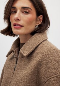 Manteau en peluche marron avec col, fermeture éclair et tissu texturé. Modèle avec des cheveux foncés à la hauteur des épaules portant des boucles d'oreilles en argent.