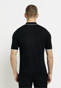 Mężczyzna z krótkimi włosami, ubrany w czarną koszulę z krótkim rękawem z białymi wykończeniami na rękawach i kołnierzyku, stojący na jednolitym jasnym tle.