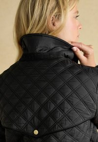 Jachetă neagră matlasată cu guler înalt, având modele în diamant, o bandă cu sigla la gât și un detaliu metalic cu clapă în spate.