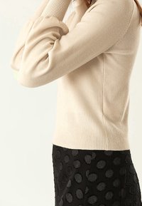 Beige Strickpullover mit geripptem Saum und puffigen Ärmeln, kombiniert mit einem schwarzen Rock mit strukturiertem Punktmuster.