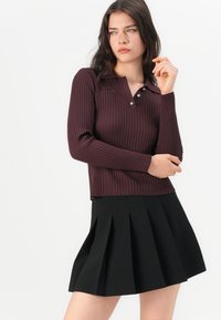 Burgundy zakrivený top s dlhými rukávmi a golierom, doplnený detailom s tromi gombíkmi, v kombinácii s čiernou plisovanou sukňou. Hladká textúra tkaniny.