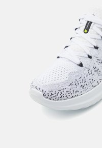 Chaussure de sport blanche avec tige en tricot respirante présentant des taches noires, bout rond et lacets blancs contrastants. Semelle blanche rembourrée.