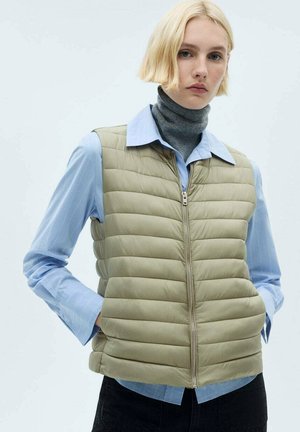 Light olive puffer vest with horizontal quilted pattern, zipper closure, worn over a blue collared shirt and a gray turtleneck.