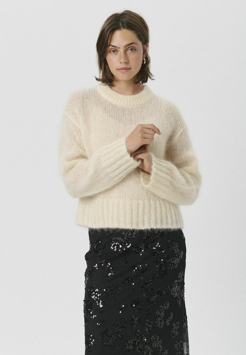 Krem pulover iz mehke pletenine z okroglim izrezom in rebrastim robom. Nošen prek črne krila z bleščicami, kar poudarja kontrast tekstur.