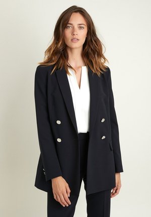 Blazer doppiopetto blu navy con dettagli in metallo argentato, vestibilità sartoriale e silhouette leggermente allungata, abbinato a un top bianco.