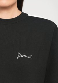 Μαύρη φούτερ μπλούζα με λευκή ρίγα στους ώμους και λευκό κεντητό λογότυπο "fiorucci" στο στήθος, φορεμένη από ένα άτομο με μακριά σκούρα μαλλιά.