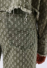 Chaqueta de mezclilla verde oliva con un patrón texturizado de pequeñas figuras blancas, bordes deshilachados y detalle de bolsillo trasero.