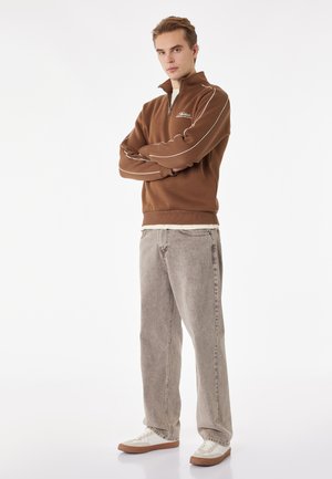 Junger Mann steht mit verschränkten Armen, trägt einen braunen Sweatshirt mit Viertel-Reißverschluss, hellgraue Jeans und weiße Sneakers vor weißem Hintergrund.