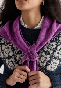 Fialový pletený pléd previazaný okolo krku, nosený cez tmavomodrý a krémový sveter so vzorom a textúrovaným pletením. Viditeľný biely volánový golier.