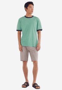 Grøn bomulds t-shirt med marineblå ribkant ved hals og ærmer, parret med beige shorts. Model med sorte sandaler, stående foran en hvid baggrund.