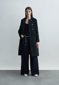 Frau steht in einem langen schwarzen Mantel, schwarzem Oberteil, weitem Hosen mit Ketten-Gürtel und weißen, spitzen Schuhen vor schlichtem grauem Hintergrund.