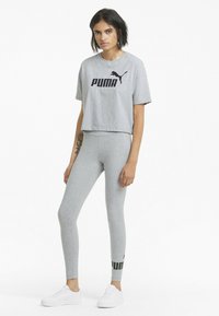 Camiseta gris recortada con el logo negro de "PUMA", combinada con leggings grises que presentan un diseño negro y verde en el tobillo, completada con zapatillas blancas.