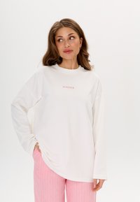 Maglietta bianca a maniche lunghe con il testo "MIRONS" rosa sul petto, caratterizzata da una vestibilità ampia e un tessuto morbido, abbinata a pantaloni a righe rosa.