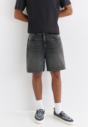 Personne portant un short en jean noir délavé, un t-shirt noir, des chaussettes blanches et des baskets noires sans lacets avec un détail de broderie blanche.