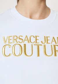 Felpa bianca con ampio testo ricamato in oro "VERSACE JEANS COUTURE" sul petto. Collo a coste e tessuto morbido.