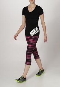 T-shirt nera con scollo a V e logo Nike bianco, abbinata a pantaloni capri a righe rosa e neri e sneakers nere con dettagli verde neon.