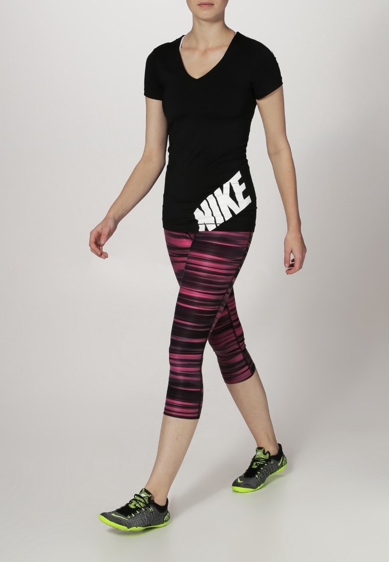 T-shirt nera con scollo a V e logo Nike bianco, abbinata a pantaloni capri a righe rosa e neri e sneakers nere con dettagli verde neon.