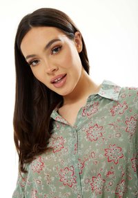 Green floral blouse featuring a button-up design, short collar, and red flower patterns. Fabric appears lightweight with a textured finish.