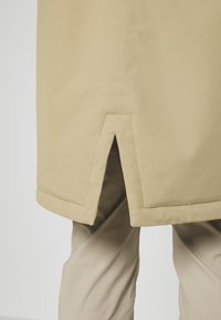 Gros plan de l'ourlet beige d’un manteau avec une fente centrale, montrant les détails de la couture et un pantalon beige dessous.