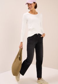 Jean noir au design ajusté, associé à une chemise blanche à manches longues. Le modèle tient un sac beige et porte une casquette pastel ainsi que des baskets.