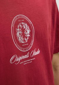 Raudonas medvilnės marškinėliai su baltu apvaliu grafiniu dizainu, kuriame pavaizduoti gėlių motyvai ir tekstas: "Sukurtas Kopenhagoje, Originals Studio, Nuo 1990 metų."