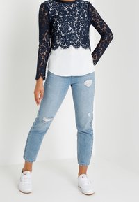 Kvinna som bär en marinblå spetströja med långa ärmar över en vit skjorta, ljusa slitna jeans och vita sneakers stående mot en enfärgad bakgrund.