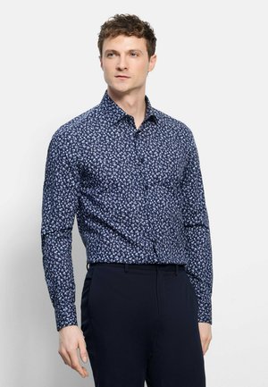 Mann trägt ein dunkelblaues, geblümtes Langarmhemd, das in dunkelblaue Hose gesteckt ist, und steht vor einem einfarbigen Hintergrund.